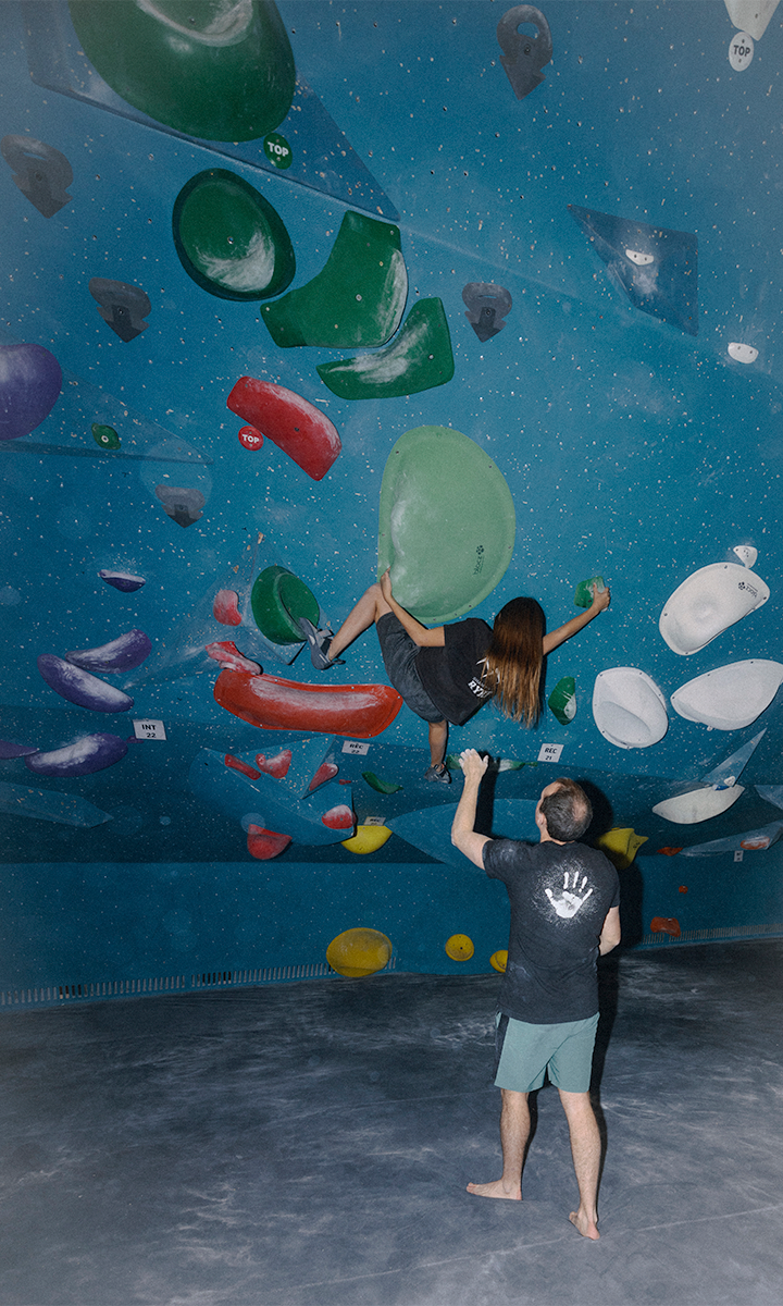 A woman climbs an indoor bouldering wall with colorful holds, while a man from Personal Climbing Coaching Austin stands below, spotting her for safety. Both wear dark shirts and shorts. The climbing wall has a blue background with star-like speckles.