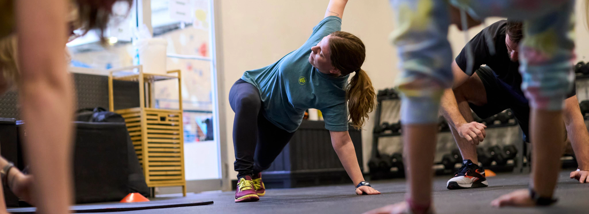 People exercising indoors, performing side stretches on the floor. In this Minneapolis gym, one person in the center wears a blue shirt and pink shoes, reaching upward, while others follow along. Personal training equipment is visible in the background.