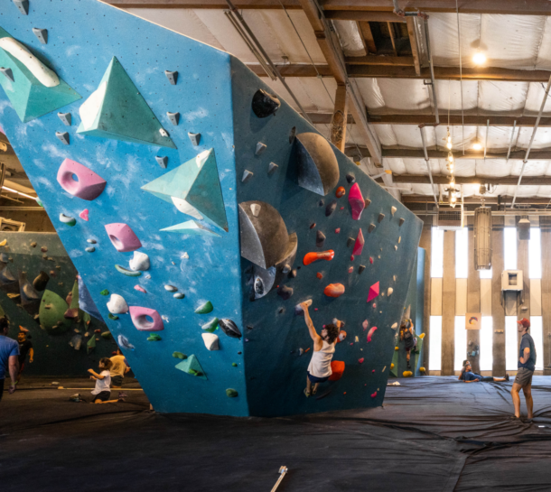 Several people are indoor rock climbing on large blue climbing walls with colorful holds in a spacious gym. Some are climbing, while others watch or rest on padded flooring. Bright lights hang from the ceiling above.