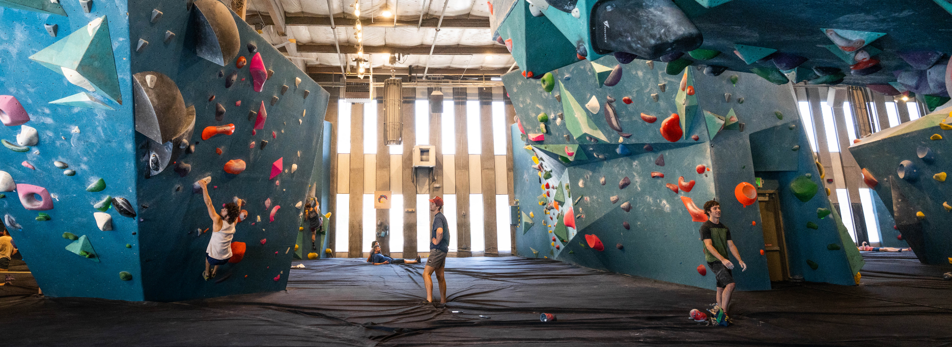 Indoor climbing gym with several people bouldering on large blue climbing walls with colorful holds. Two climbers are on the wall, two people stand on padded flooring, and bright light shines through tall windows.