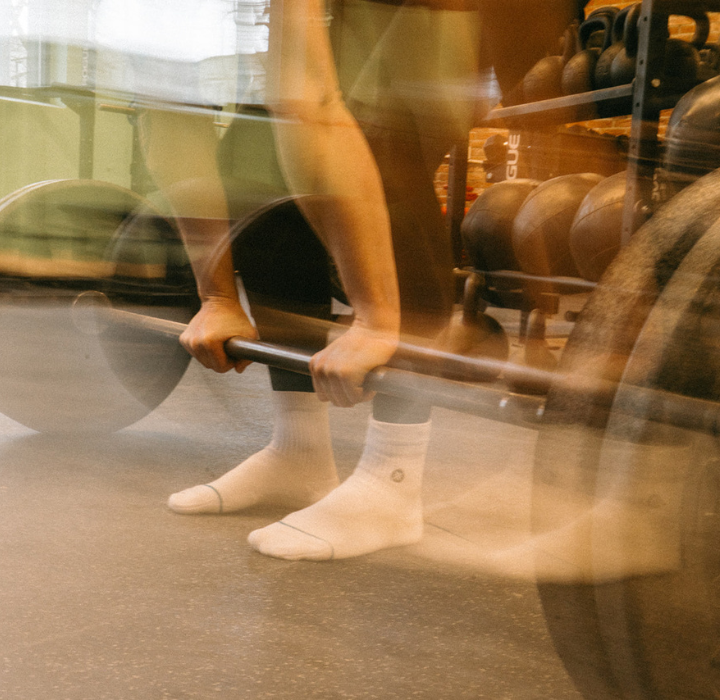 A person in white socks and black leggings lifts a barbell in a Brooklyn gym. The motion blur highlights the intensity of weightlifting, with kettlebells and hints of private instruction visible in the background.