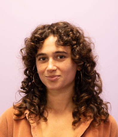 A woman with curly brown hair, wearing large hoop earrings and a peach-colored shirt, stands against a light purple background, looking at the camera with a slight smile—ready for her next adventure at an Austin Climbing Gym.