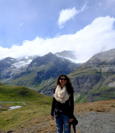 A woman in sunglasses, a white scarf, and a black top stands on a mountain path with snow-capped peaks, green slopes, and a cloudy blue sky in the background.