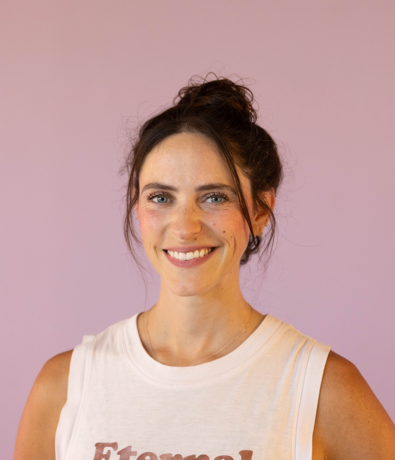 A woman with dark hair in a messy bun is smiling at the camera. She is wearing a white sleeveless top and standing in front of a plain lavender background, ready for her next adventure at an Austin climbing gym.