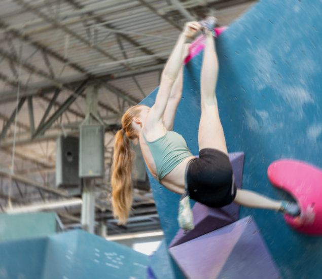 A woman with long red hair climbs an indoor bouldering wall at Austin Bouldering Climbing Gym, reaching upward with both arms and hooking a leg over a large blue hold, surrounded by geometric climbing grips.