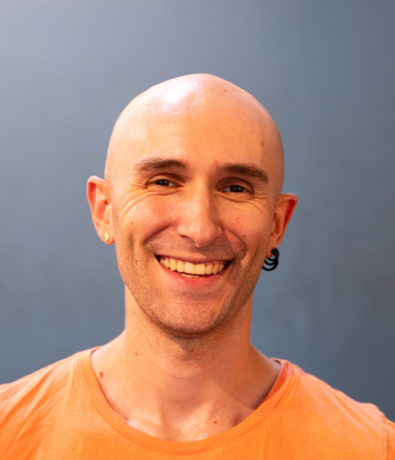 A smiling bald person with light skin, wearing a peach-colored shirt and earrings, stands in front of a plain blue-gray background, ready for another fun day at an Austin Climbing Gym.