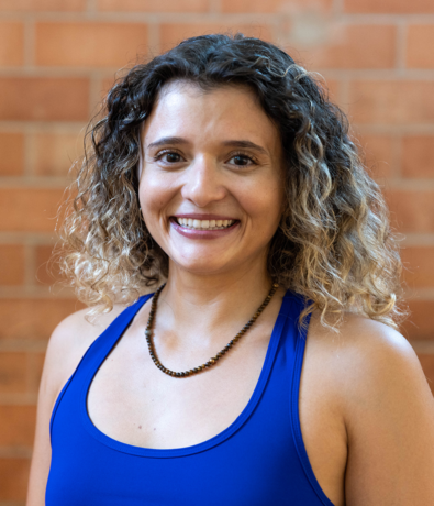 A woman with curly hair, wearing a blue sleeveless top and a beaded necklace, smiles in front of a blurred brick wall background at an Austin Climbing Gym.