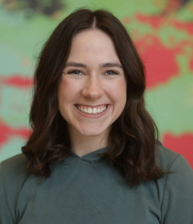 A young woman with shoulder-length brown hair and a green top smiling in front of a colorful, blurred background with green and red tones.