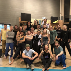A group of 21 people pose together indoors, smiling and making playful gestures. Most are barefoot and wearing casual or athletic clothing, standing in front of shelves with climbing gear. The setting appears to be a climbing gym.