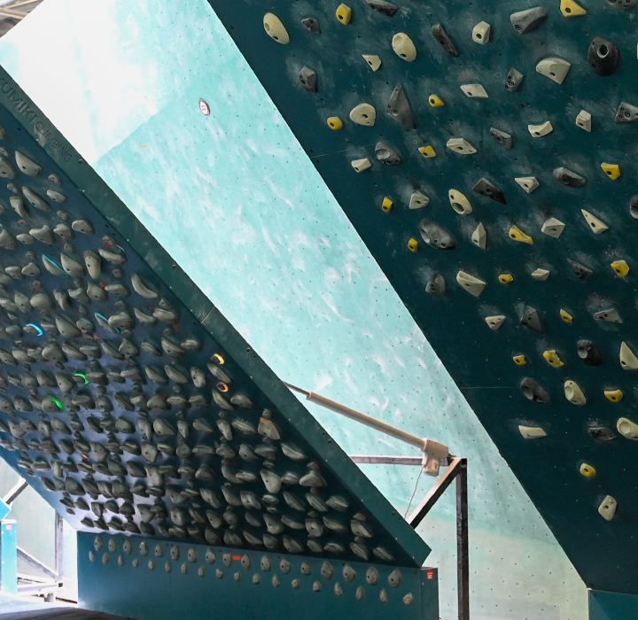 Indoor rock climbing gym in Springdale featuring three large, slanted climbing walls with colorful handholds, visible beams under a high ceiling, training boards for all levels, and seating areas in the background.