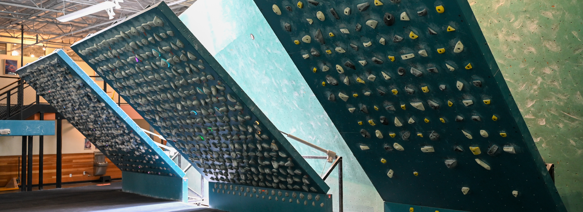 Indoor rock climbing gym in Springdale featuring three large, slanted climbing walls with colorful handholds, visible beams under a high ceiling, training boards for all levels, and seating areas in the background.