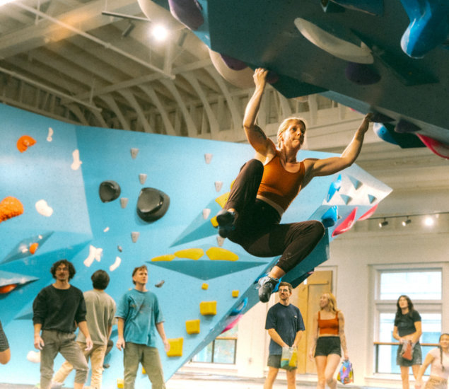 A woman in athletic wear climbs an indoor bouldering wall, gripping an overhang, while several people watch from below in the brightly lit climbing gym—an inspiring scene for New Year 2025 fitness promos.