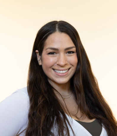 A smiling woman with long brown hair, wearing a white top and a small necklace, poses in front of a pale, softly lit background at an Austin climbing gym.