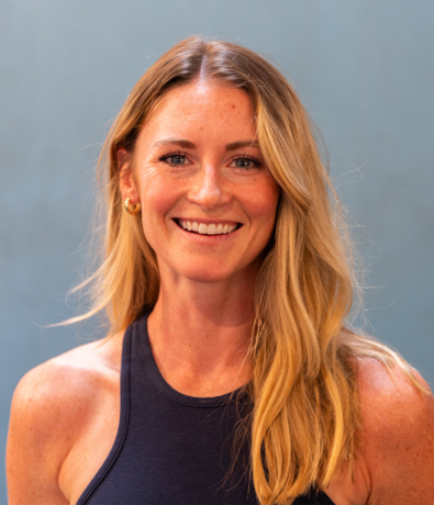 A woman with long blonde hair wearing a sleeveless dark top smiles at the camera, standing in front of a plain blue-gray background, ready for a day at an Austin Climbing Gym.