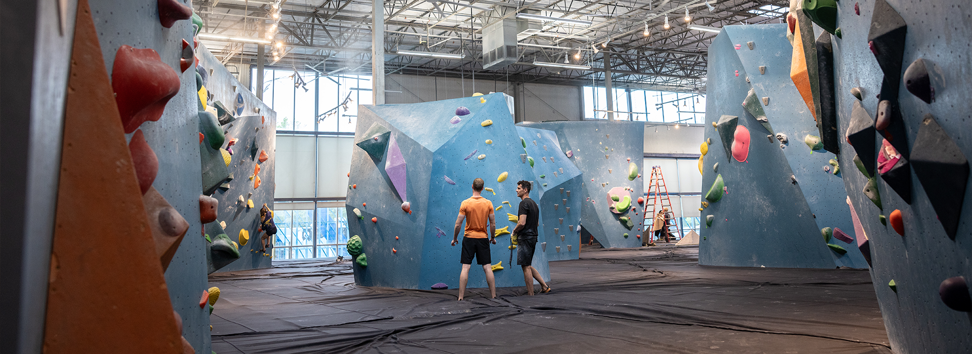 Two people in athletic clothing stand on padded floors in an Austin climbing gym, surrounded by large blue walls with colorful holds. Bright natural light streams through big windows and the ceiling above.