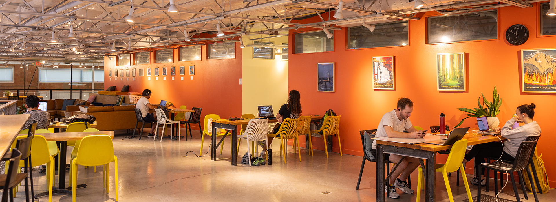 A spacious, modern workspace with people working at tables and on couches. The room has yellow chairs, an orange accent wall with framed art, large windows, and exposed ceiling beams—perfect for remote work after a session at the Austin climbing gym.