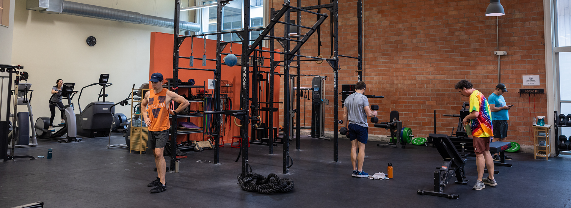 Several people work out in an Austin climbing gym with exercise equipment, weights, and fitness machines. Some use cardio machines, while others lift weights or rest. The gym features large windows and exposed brick walls.