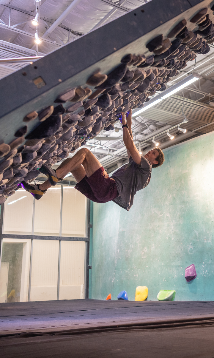 A man wearing climbing shoes, shorts, and a t-shirt is hanging from a steeply overhung wall at an Austin climbing gym, gripping holds with his hands and feet while gym mats cover the floor below.