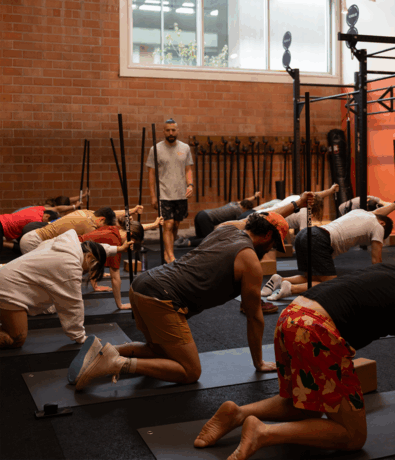 A group of people in an Austin climbing gym perform a stretching exercise on mats while an instructor observes. The room features brick walls, a large window, and gym equipment in the background.