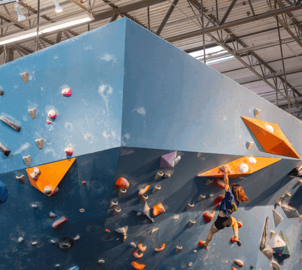 A person climbs an indoor bouldering wall with large orange holds and colorful handholds, reaching upward in a well-lit Austin climbing gym.