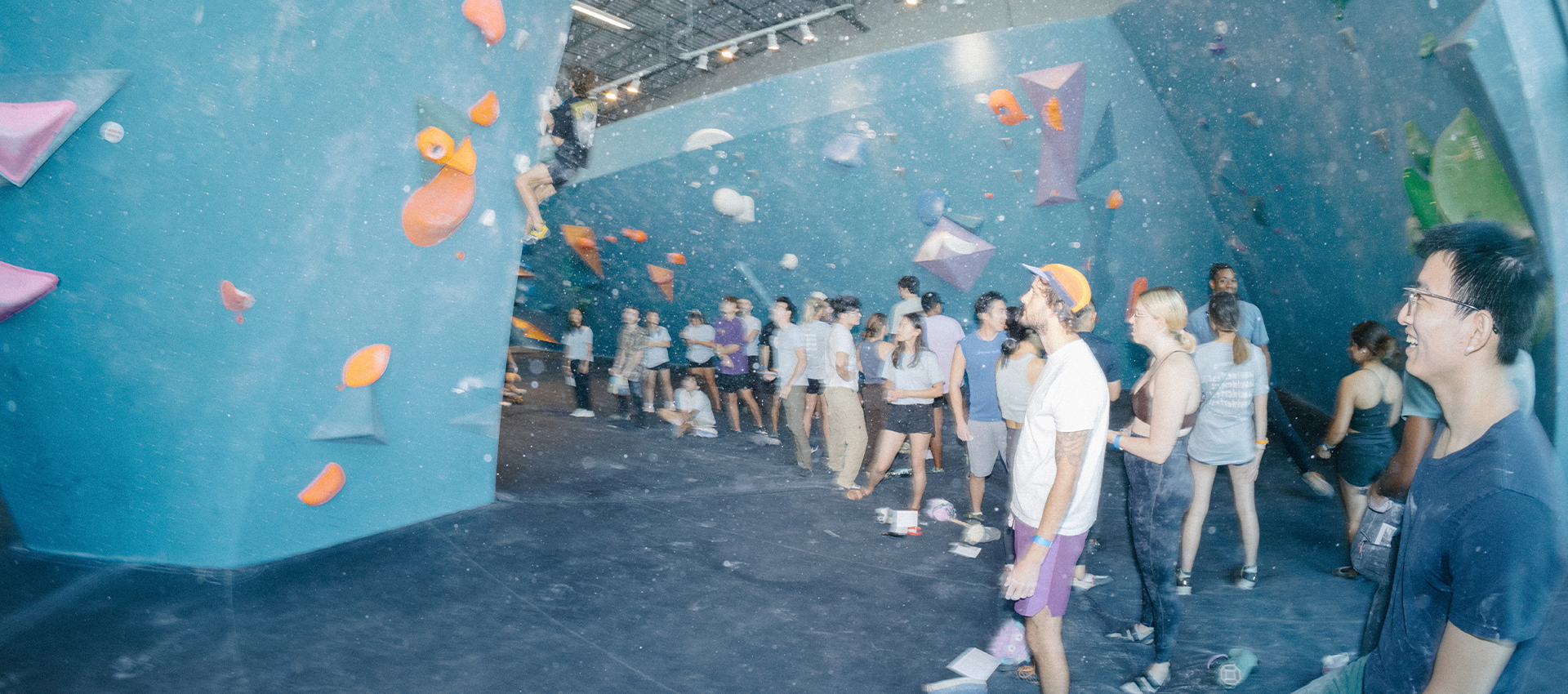 A group of people stand and watch climbers at an Austin climbing gym with blue bouldering walls and colorful holds. Some climb, while others observe and chat, creating a lively and social atmosphere.