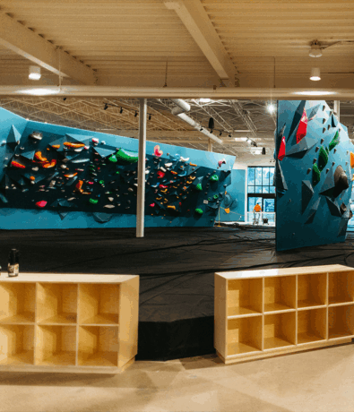 An indoor bouldering gym in Minneapolis with blue climbing walls, colorful holds, thick black mats, and wooden cubbies in the foreground. Natural light streams in through large windows—perfect for your next Bouldering Project.