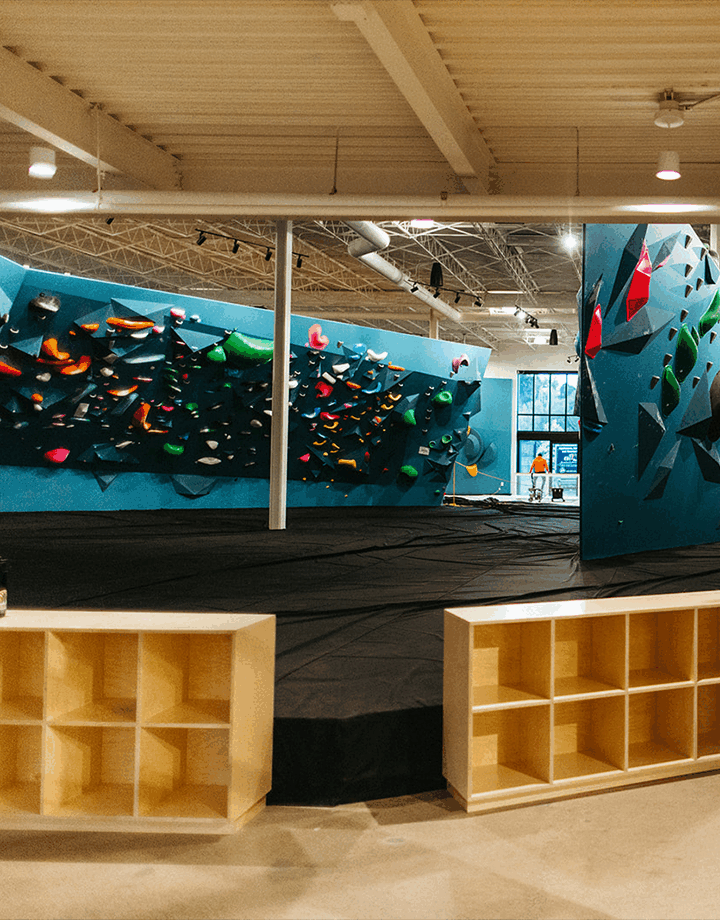 An indoor bouldering gym in Minneapolis with blue climbing walls, colorful holds, thick black mats, and wooden cubbies in the foreground. Natural light streams in through large windows—perfect for your next Bouldering Project.