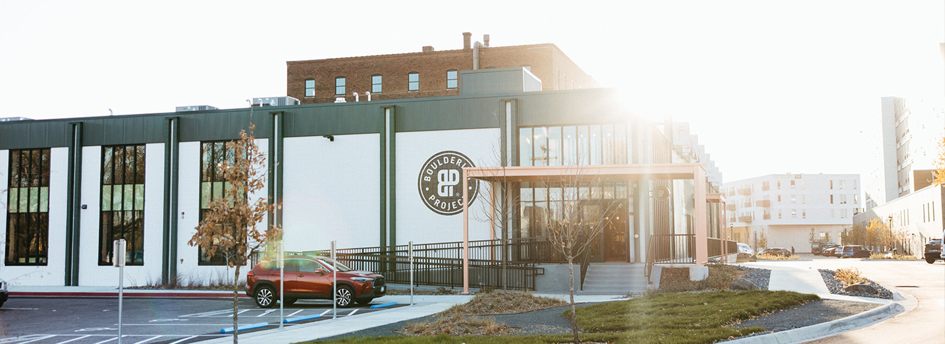 A modern building with large windows and a student choice logo on the wall, this St Paul rock climbing gym is surrounded by a parking lot with parked cars, trees, and sunlight shining brightly in the background.