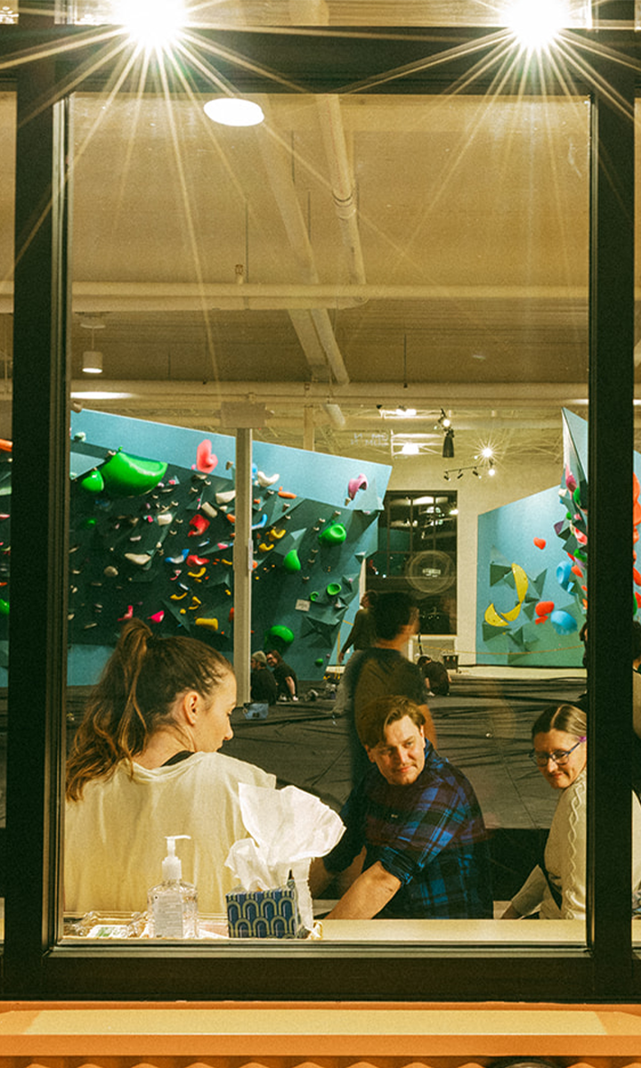 Three people sit indoors near a window at a St Paul rock climbing gym, chatting and smiling. Behind them, colorful climbing walls and climbers are visible, brightly lit. A tissue box and hand sanitizer rest on the windowsill.