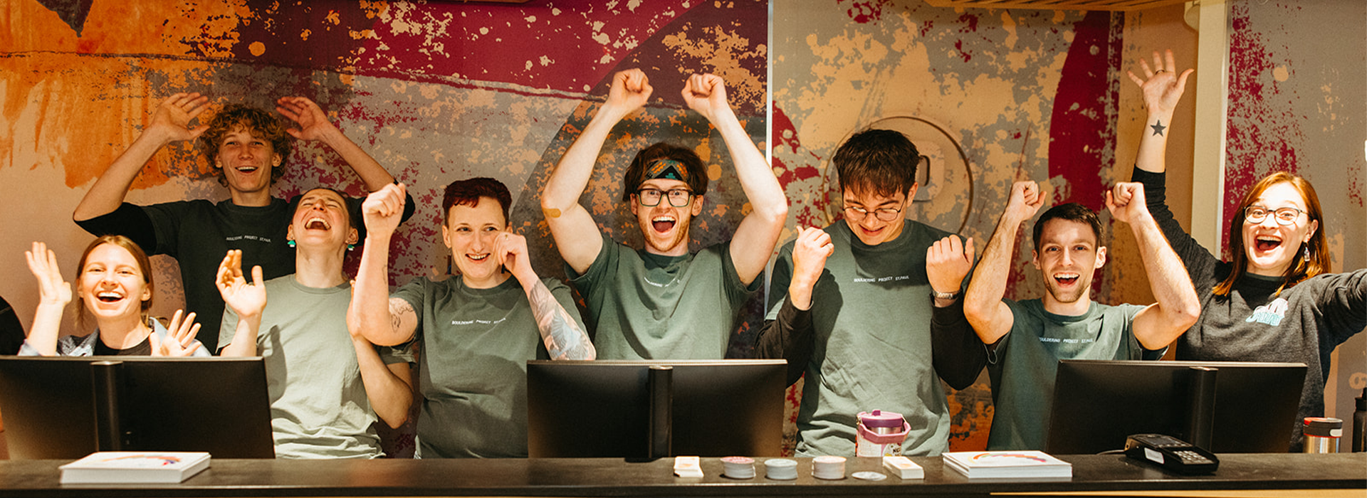 A group of eight young adults stand behind a counter at a St. Paul rock climbing gym, smiling and raising their arms in celebration. They wear matching green shirts and pose in front of a colorful, artistic wall.