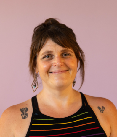 A woman with brown hair in a loose bun, wearing a black tank top with rainbow stripes, geometric earrings, and cactus tattoos on her shoulders, smiles softly against a plain light purple background—perfectly capturing the vibe of an Austin climbing gym.