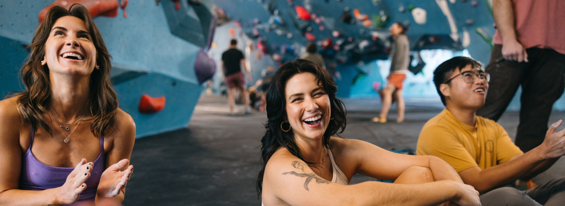 Three people smiling and laughing while sitting on the floor of Climbing Tempe, with colorful climbing walls and other climbers in the background.