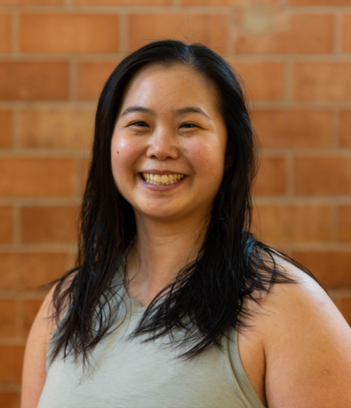 A woman with long, dark hair smiles while standing in front of a brick wall at an Austin Climbing Gym. She wears a sleeveless, light-colored top and looks directly at the camera.