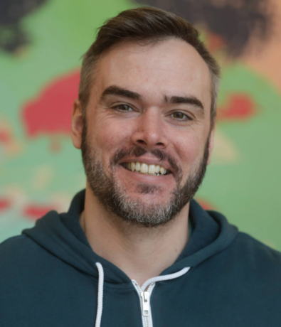 A smiling man with short brown hair and a beard, wearing a dark green hoodie with white drawstrings, stands in front of a colorful blurred background.