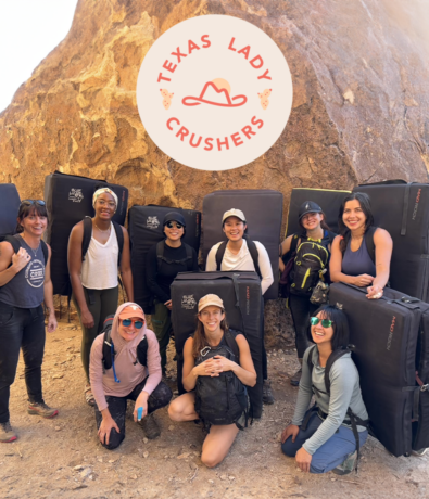 A group of eight women climbers smiling outdoors in front of large boulders, each holding crash pads. Above them is a circular logo that reads Texas Lady Crushers—part of Community & Events at Austin Bouldering Project.