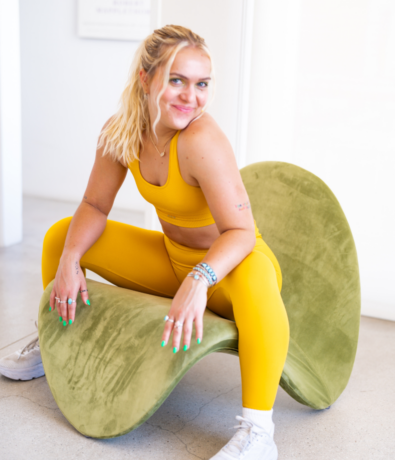 A woman in a yellow sports bra and leggings sits confidently on a modern, curved green chair, smiling at the camera. She has blonde hair in a ponytail and wears white sneakers.