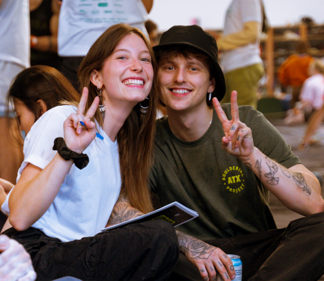 Two young adults sitting closely together in Austin, smiling at the camera and making peace signs with their hands. One holds a booklet from Height Fight; both have tattoos and casual clothing. Climbers and gear fill the background.