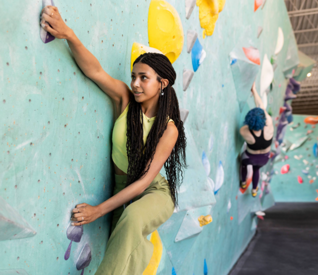A woman with long braids and a yellow-green outfit climbs an indoor rock wall during the Adult Climbing League, gripping holds with both hands; another climber is visible in the background on the colorful wall.