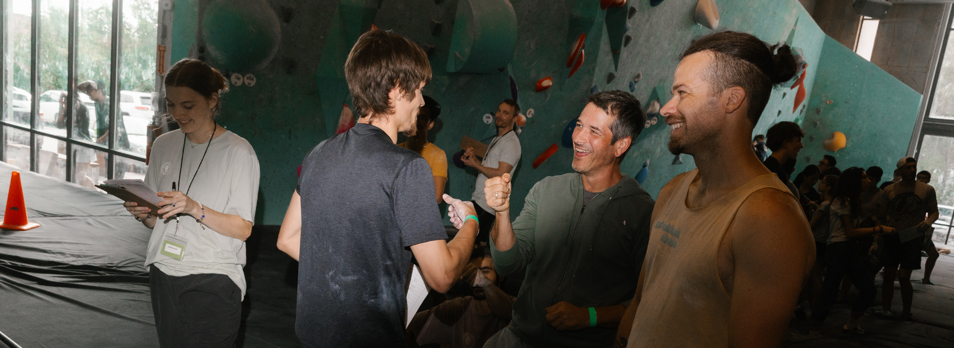 A group at an indoor rock climbing gym celebrates and smiles during Adult Climbing League, with two men giving a fist bump while others watch and a woman takes notes nearby.