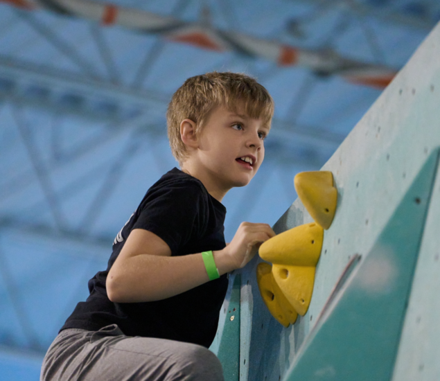 A young boy with blond hair climbs an indoor rock wall at Climb Team Minneapolis, gripping yellow handholds and looking focused. He wears a black T-shirt, gray pants, and a green wristband. The background features a blue structure.