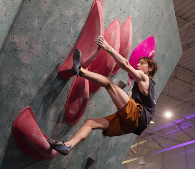 A young man wearing climbing gear scales an indoor rock climbing wall with large pink holds in Minneapolis, stretching his arms and legs to grip the surface under bright indoor lighting, representing his local Climb Team.