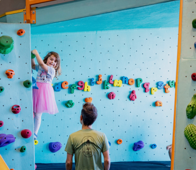 A young girl in a pink skirt smiles while climbing an indoor rock wall, as an adult watches from below. The wall, part of Minneapolis youth programs, is decorated with colorful letters of the alphabet.