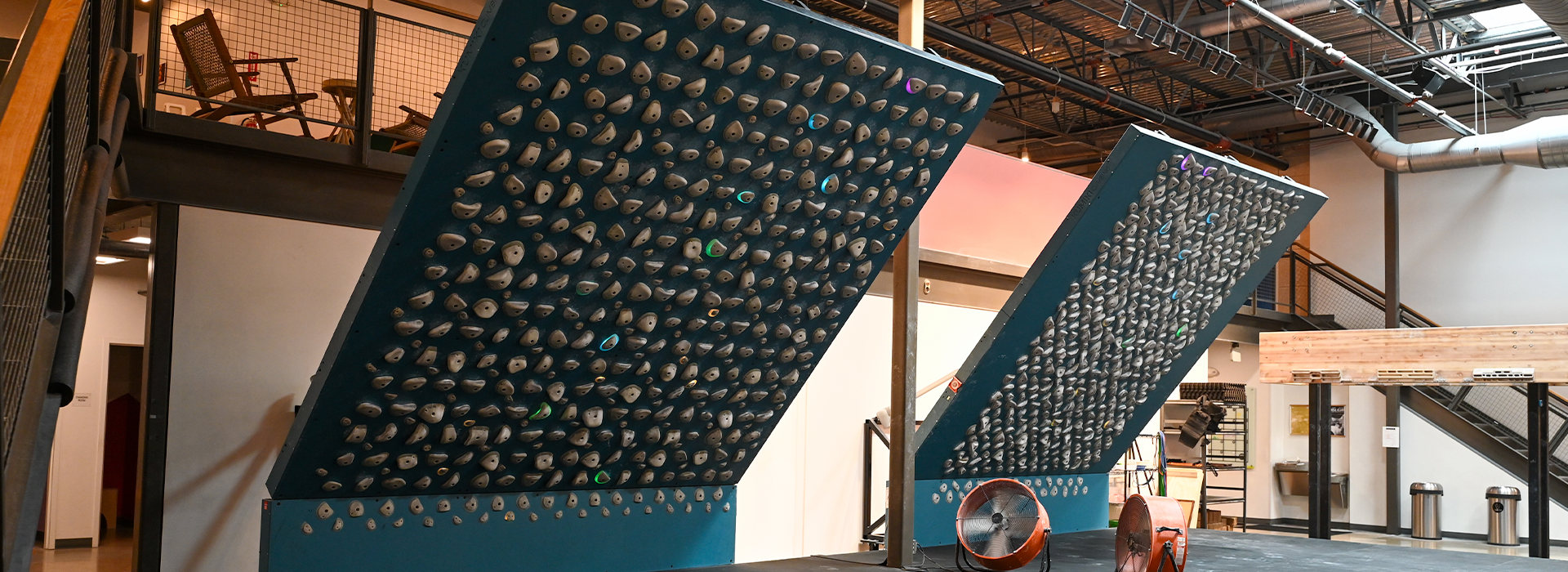 Two steeply angled Training Boards covered with numerous gray holds stand side by side in a modern climbing gym at Westgate, featuring industrial design elements and metal railings.