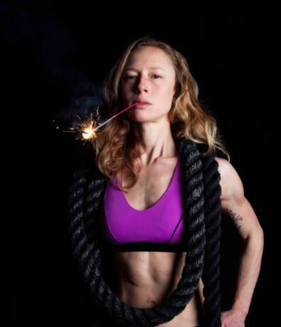 A fit woman with long blonde hair wears a purple sports bra and has a thick black rope draped over her shoulders. She holds a lit sparkler in her mouth against a black background.