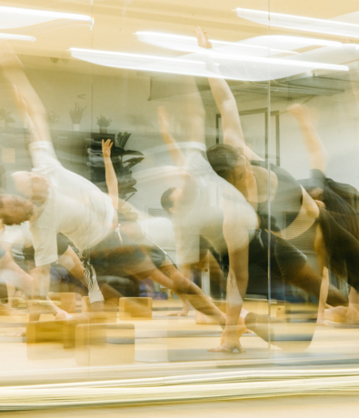 A group of people practicing yoga indoors, captured with motion blur as they stretch one arm upward in a mirrored studio. The image is dynamic, showing the fluidity of their movements.