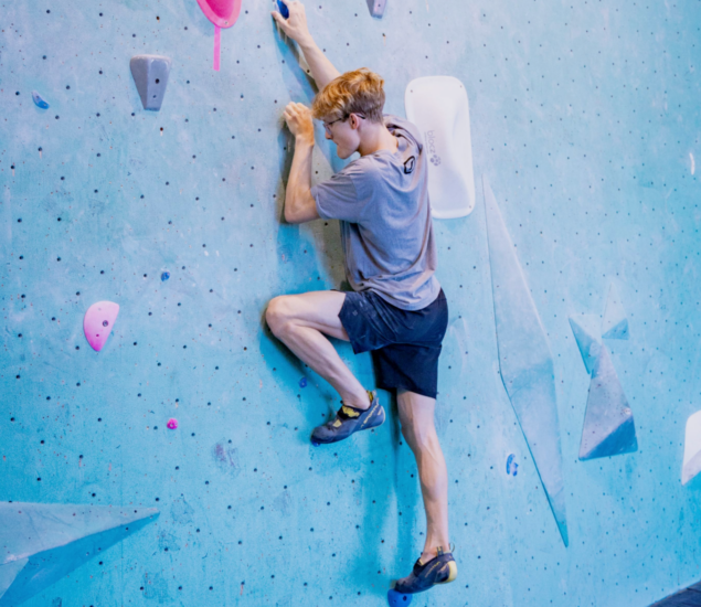 A young man wearing a gray t-shirt, black shorts, and climbing shoes is bouldering on an indoor climbing wall with blue, pink, and white holds in Minneapolis. Perfect for those interested in joining a climb team or exploring climbing locally.