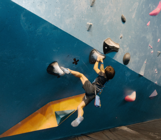 A child wearing a harness and chalk bag climbs an indoor bouldering wall in Brooklyn, gripping large black holds with both hands and reaching out with his legs—an aspiring member of local Youth Teams, suspended above padded flooring.