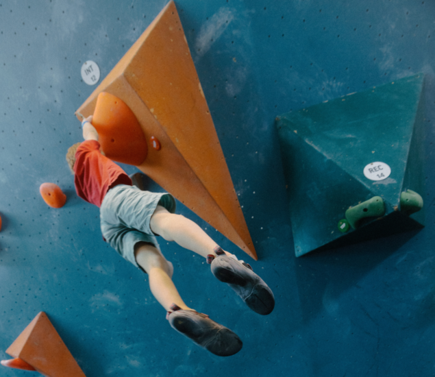 A person in a red shirt and light blue shorts climbs a blue indoor bouldering wall in Brooklyn, reaching for an orange hold on a large triangular volume. Their legs are extended behind them in mid-motion, showcasing the skill often seen in local Youth Teams.