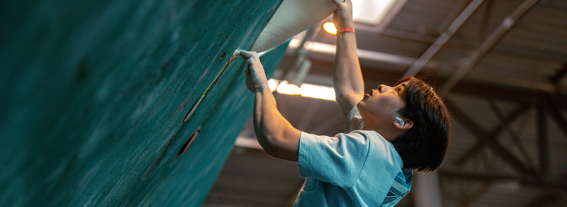 A person wearing a hearing aid climbs an indoor rock wall, gripping handholds and looking upward with focus under natural light from a skylight above—just like athletes training with Competitive Youth Climbing Teams in Austin.