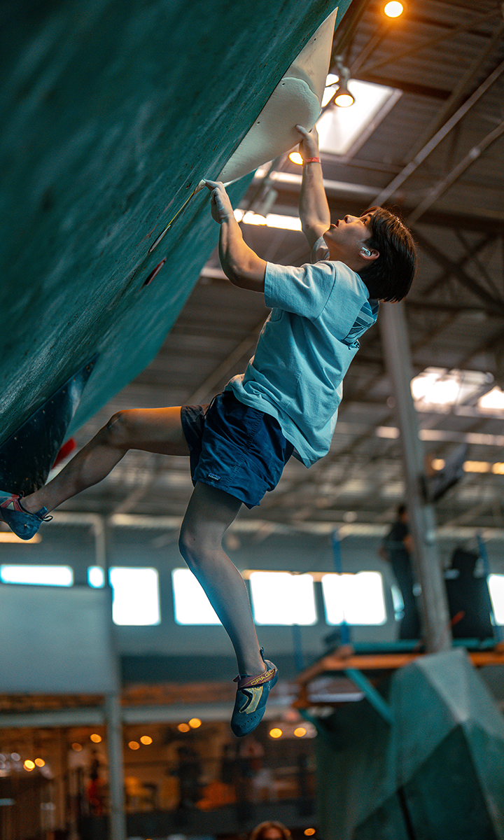 A young person wearing a light blue shirt and shorts climbs an indoor rock wall, reaching up with one hand to grip a hold—a scene typical of Competitive Youth Climbing Teams in Austin. The background shows large windows and industrial-style lighting.